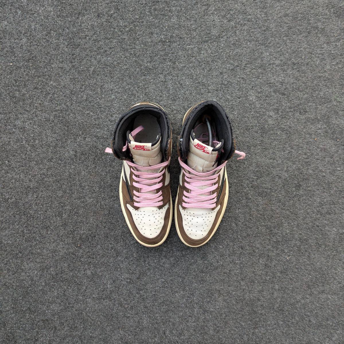 A pair of high-top sneakers viewed from above, featuring a white perforated toe box, brown leather overlays, black ankle collar, and light pink laces, placed on a textured gray surface.